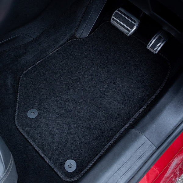 A black car floor mat installed in a vehicle's footwell, shown with the brake and accelerator pedals and part of the car interior visible.
